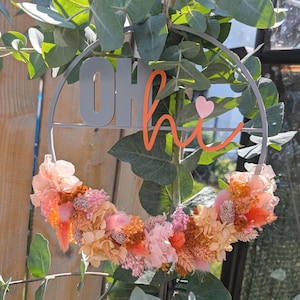 Könnte beinhalten: Ein dekorativer Metallkranz mit den Worten "OH hi" in Grau und Orange, akzentuiert mit einem kleinen rosa Herz. Der Kranz ist mit einem lebendigen Arrangement aus Trockenblumen in Pfirsich-, Rosa- und Orangetönen geschmückt, was eine einladende Ästhetik schafft.