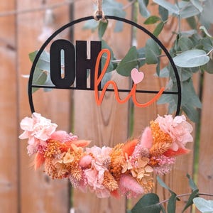 Könnte beinhalten: Ein runder schwarzer Metallring mit dem schwarzen Schriftzug OH und dem orangefarbenen kursiven Text hi sowie einem kleinen rosa Herz. Die untere Hälfte des Rings ist mit einem dichten Arrangement aus getrockneten Blumen in Rosa und Orange verziert.