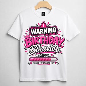 May include: A white short-sleeved t-shirt featuring a graphic design with the text WARNING BIRTHDAY BEHAVIOR LOADING... PREPARE FOR EXCESSIVE FUN! The design includes a pink crown, a loading bar, and heart accents in shades of pink and black.