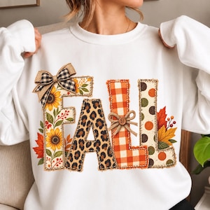 May include: A white long-sleeve sweatshirt featuring the word FALL in large block letters. The F is decorated with sunflowers and a plaid bow, the A has a leopard print, the L features orange gingham with a button, and the L has polka dots.