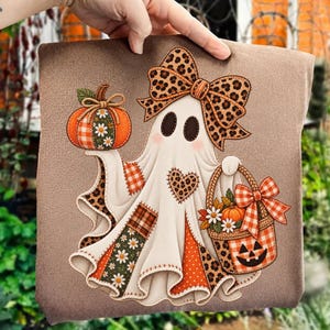 May include: A brown t-shirt featuring a graphic of a white ghost wearing a large leopard print bow. The ghost holds a pumpkin and a basket decorated with orange plaid, leopard print, and daisy patterns, with a heart patch on its chest.