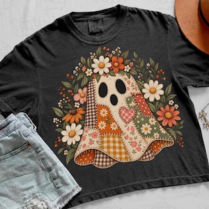 May include: A black t-shirt featuring a central graphic of a ghost made of various patchwork fabric patterns, including gingham, floral, and polka dots. The ghost is surrounded by a colorful arrangement of daisies and autumn-toned flowers.