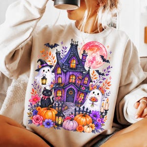 Op de afbeelding: Een beige sweater met ronde hals en een kleurrijke grafische print van een paars spookhuis, een zwarte kat, twee geesten, pompoenen, bloemen, vleermuizen en een grote roze maan, inclusief lantaarns en kaarsen in de gedetailleerde illustratie.