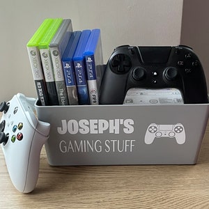 May include: A grey storage container labelled "JOSEPH'S GAMING STUFF" with a white controller icon, holds video game cases and a black controller. A white Xbox controller is beside the bin. Games for Xbox 360 and PS4 are visible.