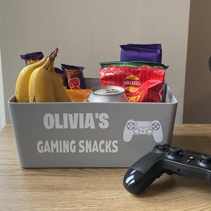 May include: A grey storage bin with the text "Olivia's Gaming Snacks" and a controller graphic. Inside are bananas, snacks, and a can. A black game controller sits on the table beside the bin.