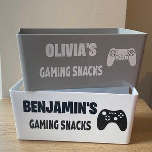 May include: Two rectangular storage bins, one grey and one white, stacked. The grey bin has the text "OLIVIA'S GAMING SNACKS" and a white game controller graphic. The white bin has the text "BENJAMIN'S GAMING SNACKS" and a black game controller graphic.