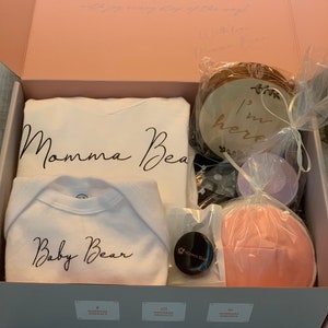 May include: A white gift box with a pink interior. The box contains a white onesie with the text "Baby Bear" in black script, a white shirt with the text "Momma Bear" in black script, a wooden sign with the text "I'm here", a small black jar with a white label that says "Momma Bear", and a pink nursing pad.