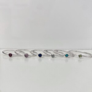 Può includere: Sette anelli d'argento con pietre preziose di diversi colori. Gli anelli sono disposti in fila da sinistra a destra. Le pietre preziose sono viola, rosa, blu, chiare, turchesi e verdi.