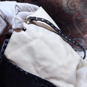 K&ouml;nnte beinhalten: Eine wei&szlig;e und schwarze Lederhandtasche mit einem Nieten-Design. Die Tasche hat einen schwarzen Riemen mit silberfarbener Hardware und schwarzen Nieten. Die Tasche wird von einer behandschuhten Hand gehalten.