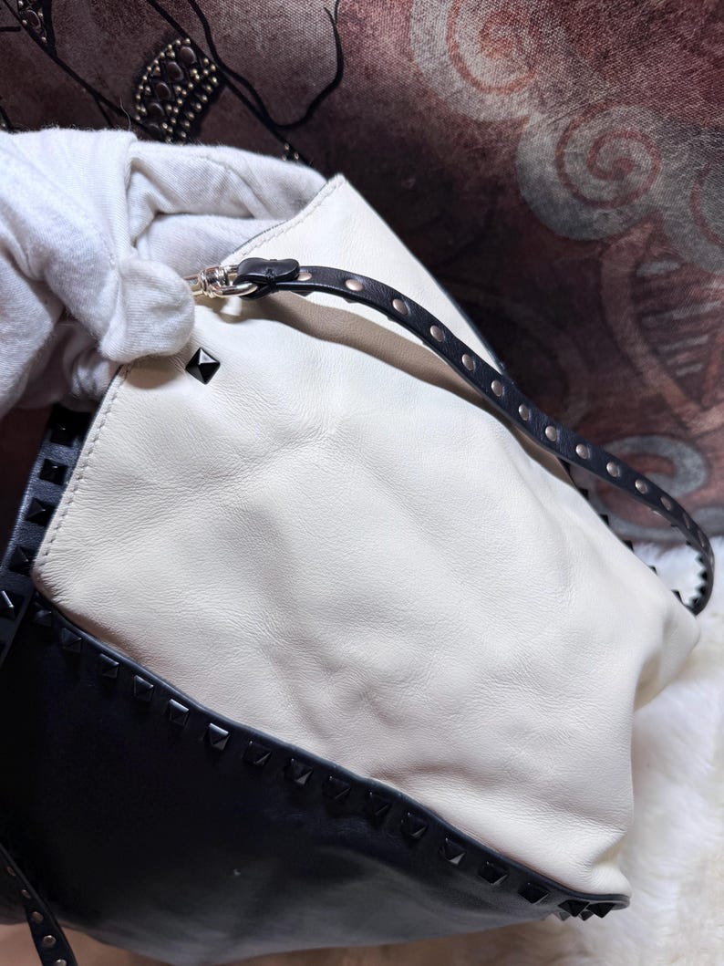 K&ouml;nnte beinhalten: Eine wei&szlig;e und schwarze Lederhandtasche mit einem Nieten-Design. Die Tasche hat einen schwarzen Riemen mit silberfarbener Hardware und schwarzen Nieten. Die Tasche wird von einer behandschuhten Hand gehalten.
