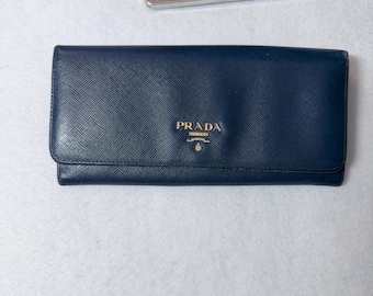 May include: A navy blue leather wallet with a fold-over flap. The gold "PRADA" logo is centered on the front. The wallet is rectangular and appears to be closed.