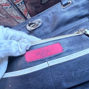 K&ouml;nnte beinhalten: Nahaufnahme einer schwarzen Lederhandtasche mit silberfarbenen Beschl&auml;gen. Der Innenraum zeigt ein pinkfarbenes Etikett mit dem Logo "Valentino Garavani". Eine wei&szlig;e, behandschuhte Hand ist teilweise sichtbar.