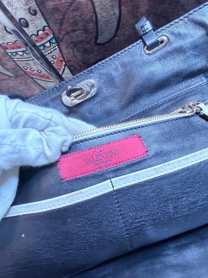 K&ouml;nnte beinhalten: Nahaufnahme einer schwarzen Lederhandtasche mit silberfarbenen Beschl&auml;gen. Der Innenraum zeigt ein pinkfarbenes Etikett mit dem Logo "Valentino Garavani". Eine wei&szlig;e, behandschuhte Hand ist teilweise sichtbar.