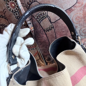 May include: A beige canvas handbag featuring a prominent pink and black striped pattern. The bag includes a black leather handle attached with silver-toned metal hardware, a black interior lining, and a structured opening with visible stitching details.