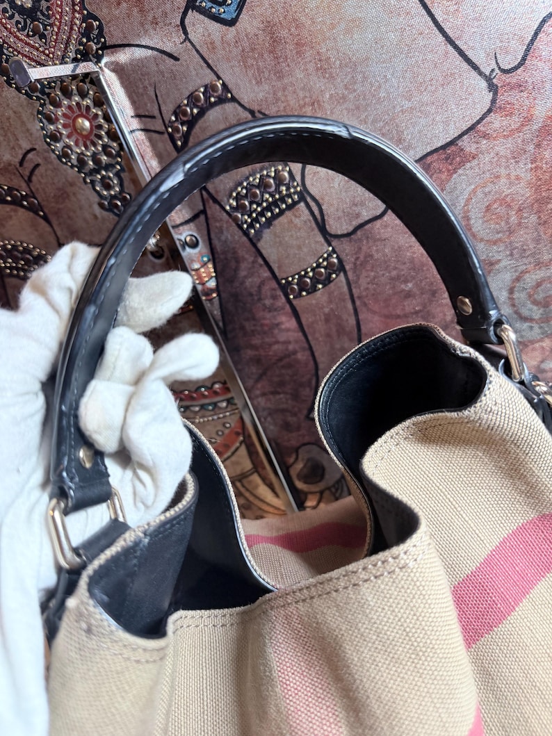 May include: A beige canvas handbag featuring a prominent pink and black striped pattern. The bag includes a black leather handle attached with silver-toned metal hardware, a black interior lining, and a structured opening with visible stitching details.