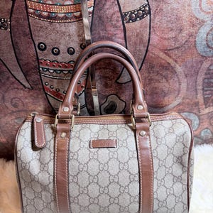 May include: A beige and brown Gucci handbag featuring the signature GG monogram pattern, brown leather trim, dual top handles, and a top zipper closure. The bag has a rectangular shape with rounded corners and a small rectangular logo patch on the front.