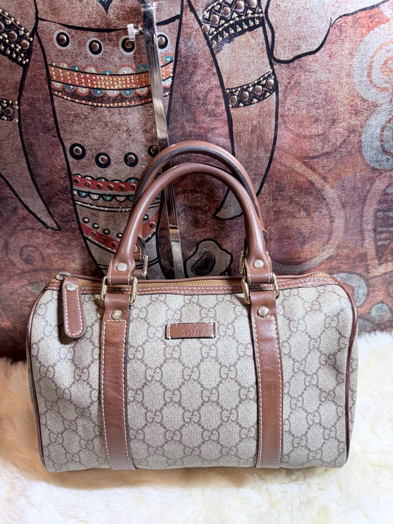 May include: A beige and brown Gucci handbag featuring the signature GG monogram pattern, brown leather trim, dual top handles, and a top zipper closure. The bag has a rectangular shape with rounded corners and a small rectangular logo patch on the front.