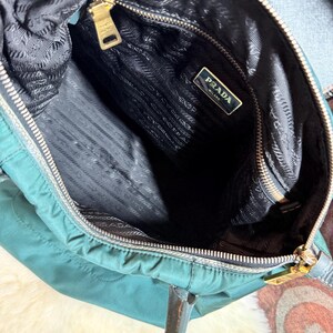 K&ouml;nnte beinhalten: Offene Ansicht einer tealfarbenen Prada-Handtasche. Der Innenraum ist schwarz mit einem Prada-Etikett. Die Tasche hat einen goldenen Rei&szlig;verschluss und goldfarbene Beschl&auml;ge. Die Griffe sind braun.