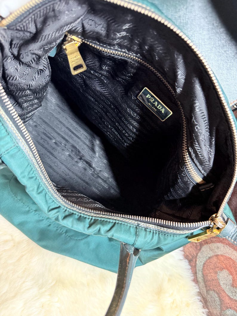 K&ouml;nnte beinhalten: Offene Ansicht einer tealfarbenen Prada-Handtasche. Der Innenraum ist schwarz mit einem Prada-Etikett. Die Tasche hat einen goldenen Rei&szlig;verschluss und goldfarbene Beschl&auml;ge. Die Griffe sind braun.