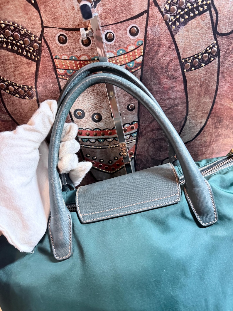 K&ouml;nnte beinhalten: T&uuml;rkisfarbene Handtasche mit passenden Griffen und einer kleinen, rechteckigen Klappe. Die Griffe sind gebogen und gen&auml;ht, und die Tasche scheint aus einem glatten Material, m&ouml;glicherweise Nylon, zu bestehen. Die Tasche hat einen Rei&szlig;verschluss.