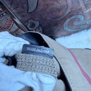 May include: A beige canvas bag featuring a red and black striped pattern with a black leather interior tag that clearly displays the text MADE IN CHINA in white capital letters. The bag has a textured fabric exterior and dark leather trim details.