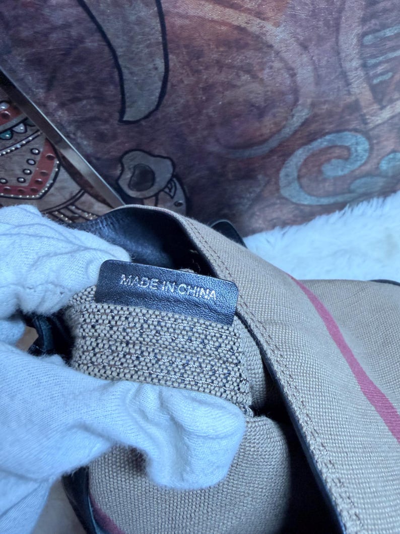 May include: A beige canvas bag featuring a red and black striped pattern with a black leather interior tag that clearly displays the text MADE IN CHINA in white capital letters. The bag has a textured fabric exterior and dark leather trim details.