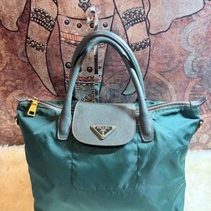 K&ouml;nnte beinhalten: Eine tealfarbene Prada Handtasche mit einem oberen Griff und einem Klappverschluss. Die Tasche verf&uuml;gt &uuml;ber goldfarbene Hardware, ein kleines Prada-Logo und einen Rei&szlig;verschluss. Die Tasche ist aus Nylon gefertigt.