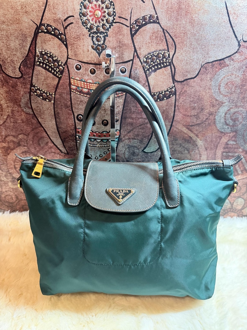 K&ouml;nnte beinhalten: Eine tealfarbene Prada Handtasche mit einem oberen Griff und einem Klappverschluss. Die Tasche verf&uuml;gt &uuml;ber goldfarbene Hardware, ein kleines Prada-Logo und einen Rei&szlig;verschluss. Die Tasche ist aus Nylon gefertigt.