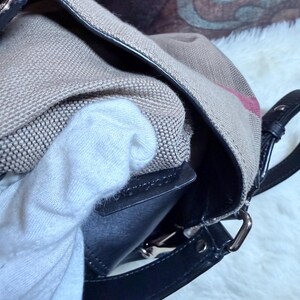 May include: A beige canvas handbag featuring a signature plaid pattern with black leather trim and a black leather strap attachment. The bag has a structured black leather interior edge and silver-toned metal hardware connecting the strap.