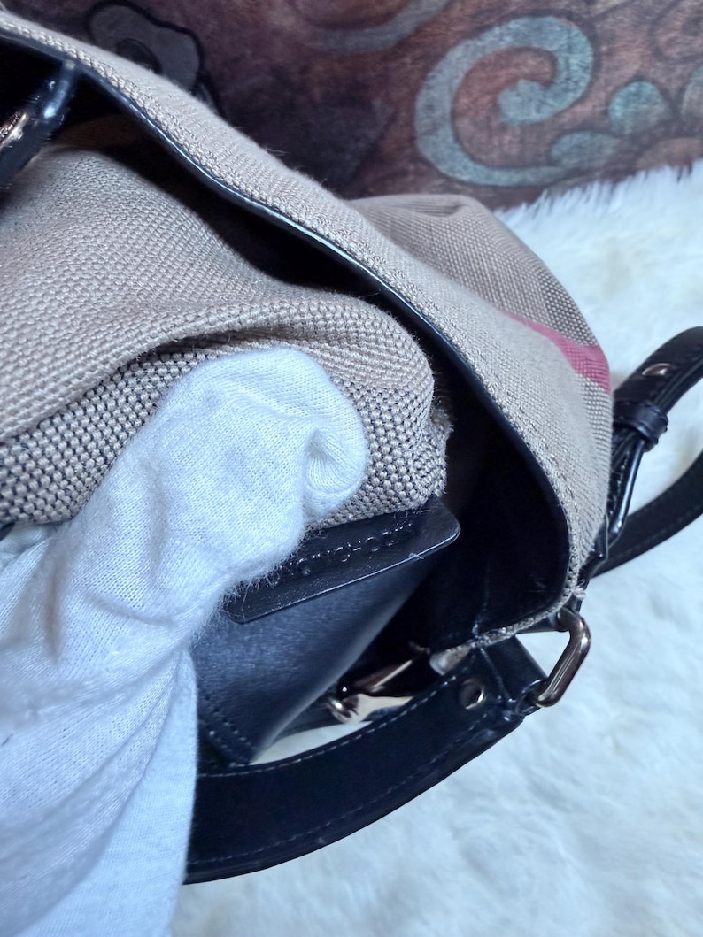May include: A beige canvas handbag featuring a signature plaid pattern with black leather trim and a black leather strap attachment. The bag has a structured black leather interior edge and silver-toned metal hardware connecting the strap.