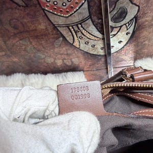May include: A brown leather interior tag inside a bag featuring the embossed serial number 193603 001998. The tag is attached to a brown leather trim edge, positioned above a grey fabric lining, with a metallic zipper pull visible nearby.
