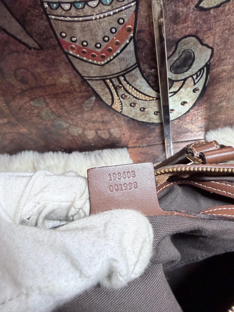 May include: A brown leather interior tag inside a bag featuring the embossed serial number 193603 001998. The tag is attached to a brown leather trim edge, positioned above a grey fabric lining, with a metallic zipper pull visible nearby.