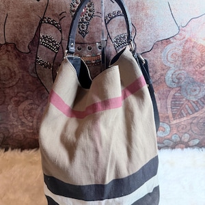 May include: A bucket bag made of beige canvas featuring a prominent plaid pattern in black, white, and red stripes. The bag includes a black leather handle and a black leather shoulder strap, with a structured base and a wide, open top opening.