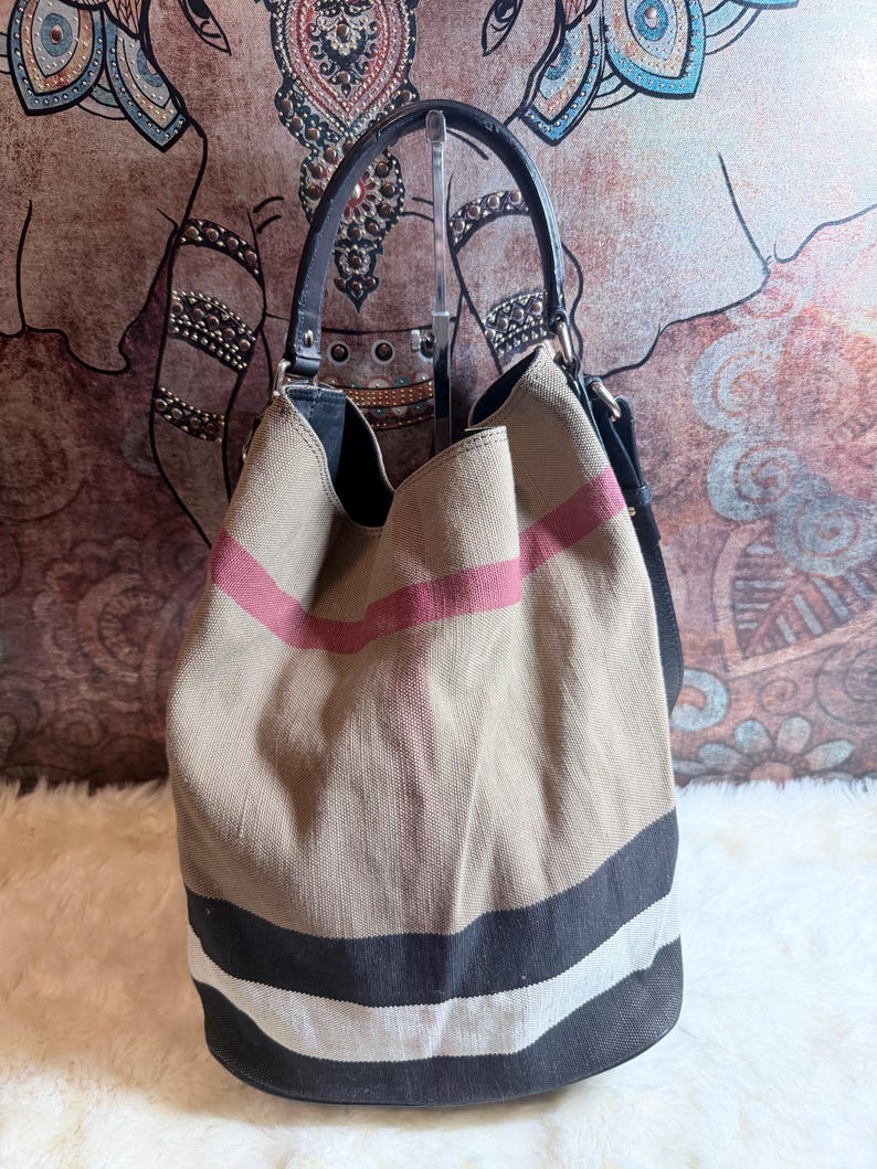 May include: A bucket bag made of beige canvas featuring a prominent plaid pattern in black, white, and red stripes. The bag includes a black leather handle and a black leather shoulder strap, with a structured base and a wide, open top opening.