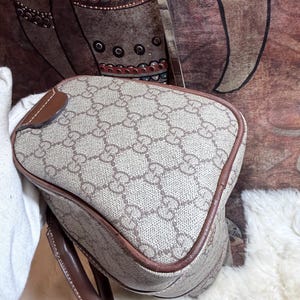May include: A beige and brown handbag featuring the iconic interlocking GG monogram pattern across the exterior. The bag is constructed with a structured shape, brown leather piping along the edges, and a matching brown leather handle attached to the side.