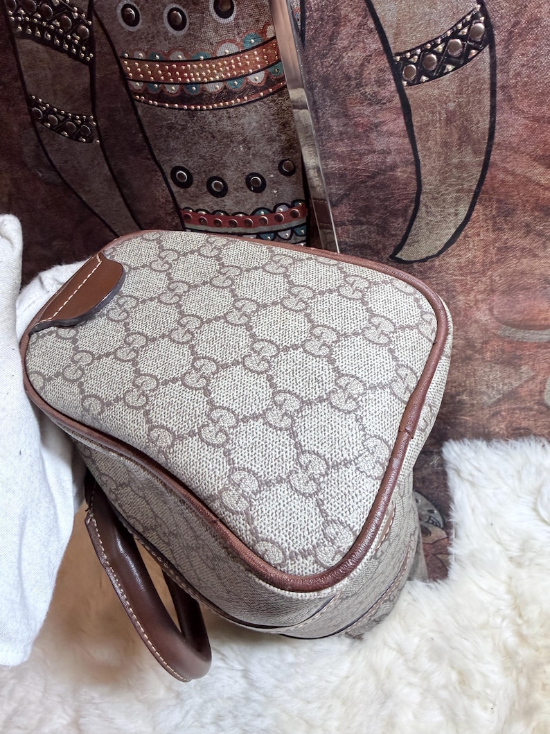 May include: A beige and brown handbag featuring the iconic interlocking GG monogram pattern across the exterior. The bag is constructed with a structured shape, brown leather piping along the edges, and a matching brown leather handle attached to the side.