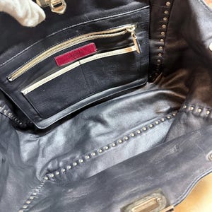K&ouml;nnte beinhalten: Schwarze Leder-Tragetasche mit silberfarbenen Nieten. Der Innenraum verf&uuml;gt &uuml;ber eine Rei&szlig;verschlusstasche mit einem roten Valentino-Etikett. Die Tasche hat einen silbernen Verschluss und eine Rei&szlig;verschlusstasche.
