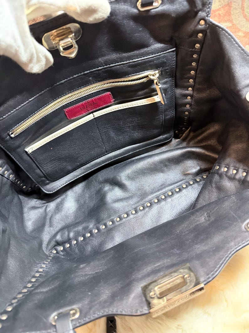 K&ouml;nnte beinhalten: Schwarze Leder-Tragetasche mit silberfarbenen Nieten. Der Innenraum verf&uuml;gt &uuml;ber eine Rei&szlig;verschlusstasche mit einem roten Valentino-Etikett. Die Tasche hat einen silbernen Verschluss und eine Rei&szlig;verschlusstasche.