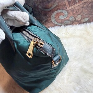 K&ouml;nnte beinhalten: Dunkelgr&uuml;ne Handtasche mit goldenem Rei&szlig;verschluss und goldenem Metallanh&auml;nger mit der Aufschrift "PRADA MILANO". Die Tasche hat einen dunkelgr&uuml;nen Griff und einen silbernen Metallring. Die Tasche ist aus glattem, gl&auml;nzendem Material.
