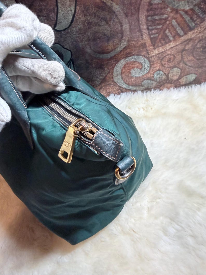 K&ouml;nnte beinhalten: Dunkelgr&uuml;ne Handtasche mit goldenem Rei&szlig;verschluss und goldenem Metallanh&auml;nger mit der Aufschrift "PRADA MILANO". Die Tasche hat einen dunkelgr&uuml;nen Griff und einen silbernen Metallring. Die Tasche ist aus glattem, gl&auml;nzendem Material.