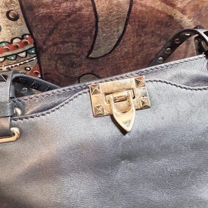 K&ouml;nnte beinhalten: Nahaufnahme einer dunkelgrauen Lederhandtasche. Die Tasche hat einen goldfarbenen Verschluss mit spitzem Ende und ist mit kleinen, quadratischen goldenen Nieten verziert. Der Riemen ist oben mit silbernen Nieten versehen.