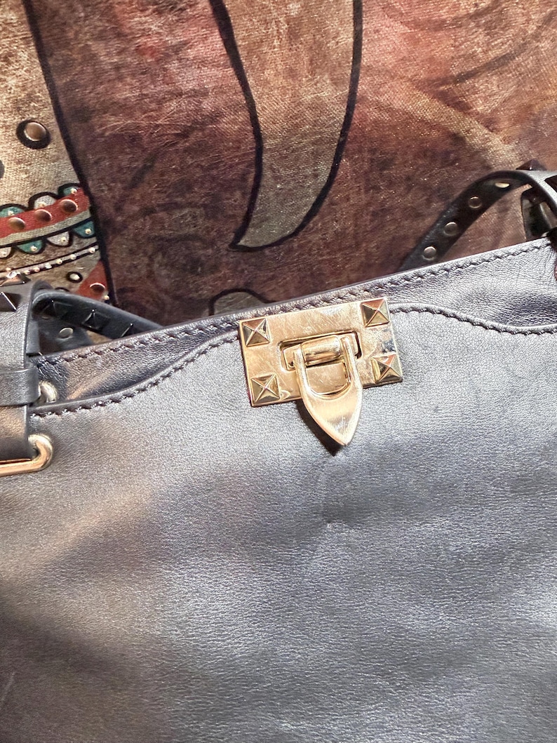K&ouml;nnte beinhalten: Nahaufnahme einer dunkelgrauen Lederhandtasche. Die Tasche hat einen goldfarbenen Verschluss mit spitzem Ende und ist mit kleinen, quadratischen goldenen Nieten verziert. Der Riemen ist oben mit silbernen Nieten versehen.
