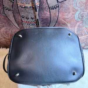 May include: A black handbag base featuring a smooth, dark surface with four small, silver-toned metal feet positioned at each corner to provide stability. The rectangular bottom has rounded edges and shows subtle signs of surface wear.