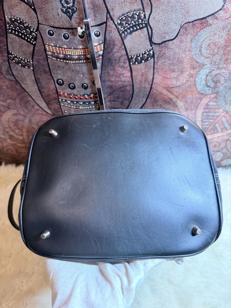 May include: A black handbag base featuring a smooth, dark surface with four small, silver-toned metal feet positioned at each corner to provide stability. The rectangular bottom has rounded edges and shows subtle signs of surface wear.