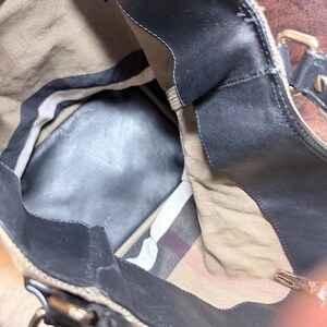 May include: A tote bag with a black leather trim and a tan canvas interior. The bag features a classic beige, black, and red plaid pattern lining on the bottom section, black leather handles, and metallic hardware attachments for the straps.