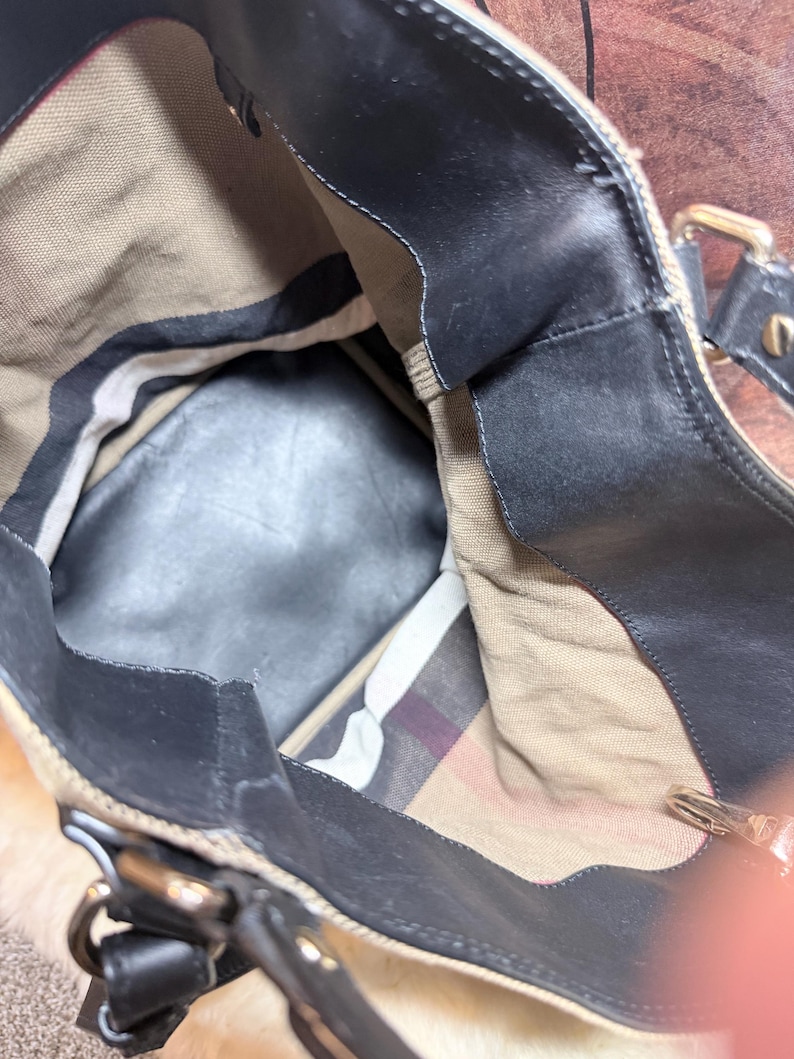 May include: A tote bag with a black leather trim and a tan canvas interior. The bag features a classic beige, black, and red plaid pattern lining on the bottom section, black leather handles, and metallic hardware attachments for the straps.