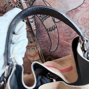 May include: A beige canvas handbag featuring a prominent red and black striped pattern. The bag includes a dark black leather handle attached with silver-toned metal hardware. The interior is lined with black fabric and features a structured opening.