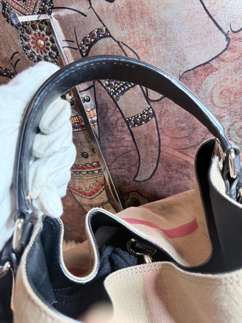 May include: A beige canvas handbag featuring a prominent red and black striped pattern. The bag includes a dark black leather handle attached with silver-toned metal hardware. The interior is lined with black fabric and features a structured opening.
