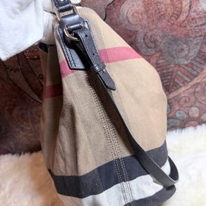 May include: A beige canvas hobo bag featuring a signature check pattern in black, white, and red stripes. The bag includes a black leather shoulder strap attached with silver-toned metal hardware, including a buckle detail on the side strap.