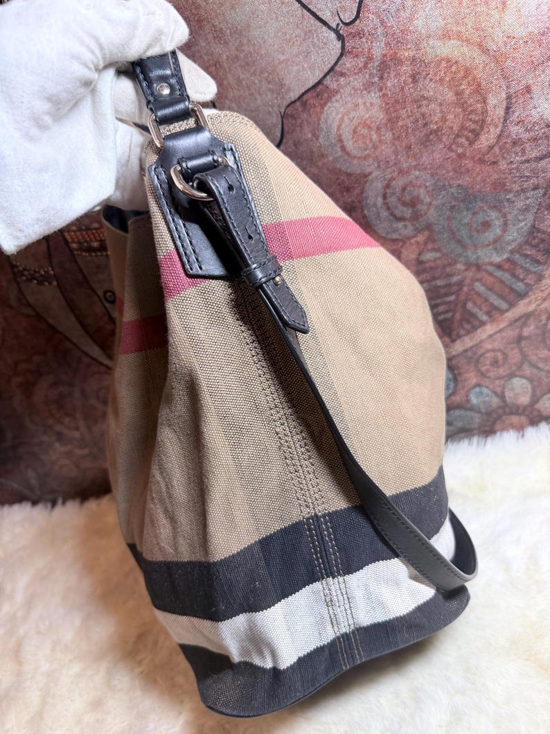 May include: A beige canvas hobo bag featuring a signature check pattern in black, white, and red stripes. The bag includes a black leather shoulder strap attached with silver-toned metal hardware, including a buckle detail on the side strap.